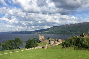 Das Foto zeigt die Ruinen von Urquhart Castle, einer mittelalterlichen Burg am Ufer des berühmten schottischen Sees Loch Ness. Die Szene spielt sich an einem sonnigen Tag mit vereinzelten Wolken am Himmel ab. Im Vordergrund befindet sich eine weitläufige grüne Wiese, begrenzt durch einen hölzernen Zaun. Einige Besucher sind auf dem Weg, der zur Burgruine hinabführt, unterwegs. Die Burganlage selbst besteht aus mehreren teils verfallenen Mauern und einem gut erhaltenen Turm, der auf einem kleinen Hügel direkt am Wasser steht. Im Hintergrund erstreckt sich der tiefblaue See bis zu den bewaldeten Hügeln am anderen Ufer. Die Landschaft vermittelt eine friedliche, historische Atmosphäre.